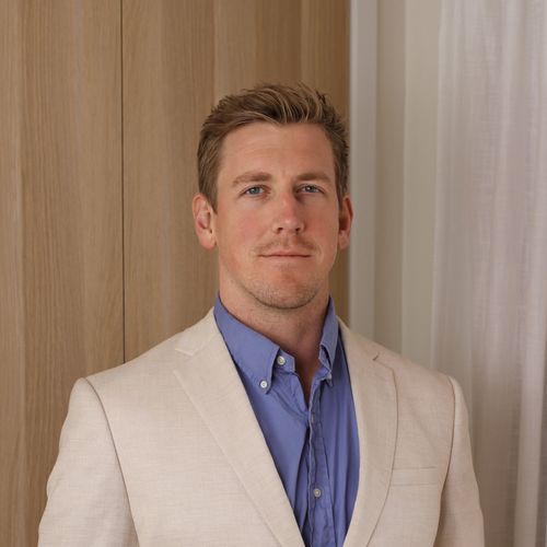 Portrait of a man with light brown hair wearing a beige blazer and blue shirt standing in front of wooden panel and white curtain.