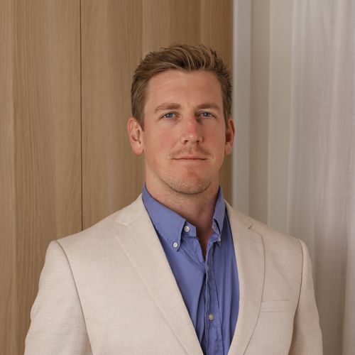 Portrait of a man with short light brown hair wearing a beige blazer and blue shirt, standing indoors against a wooden panel and white curtain background.