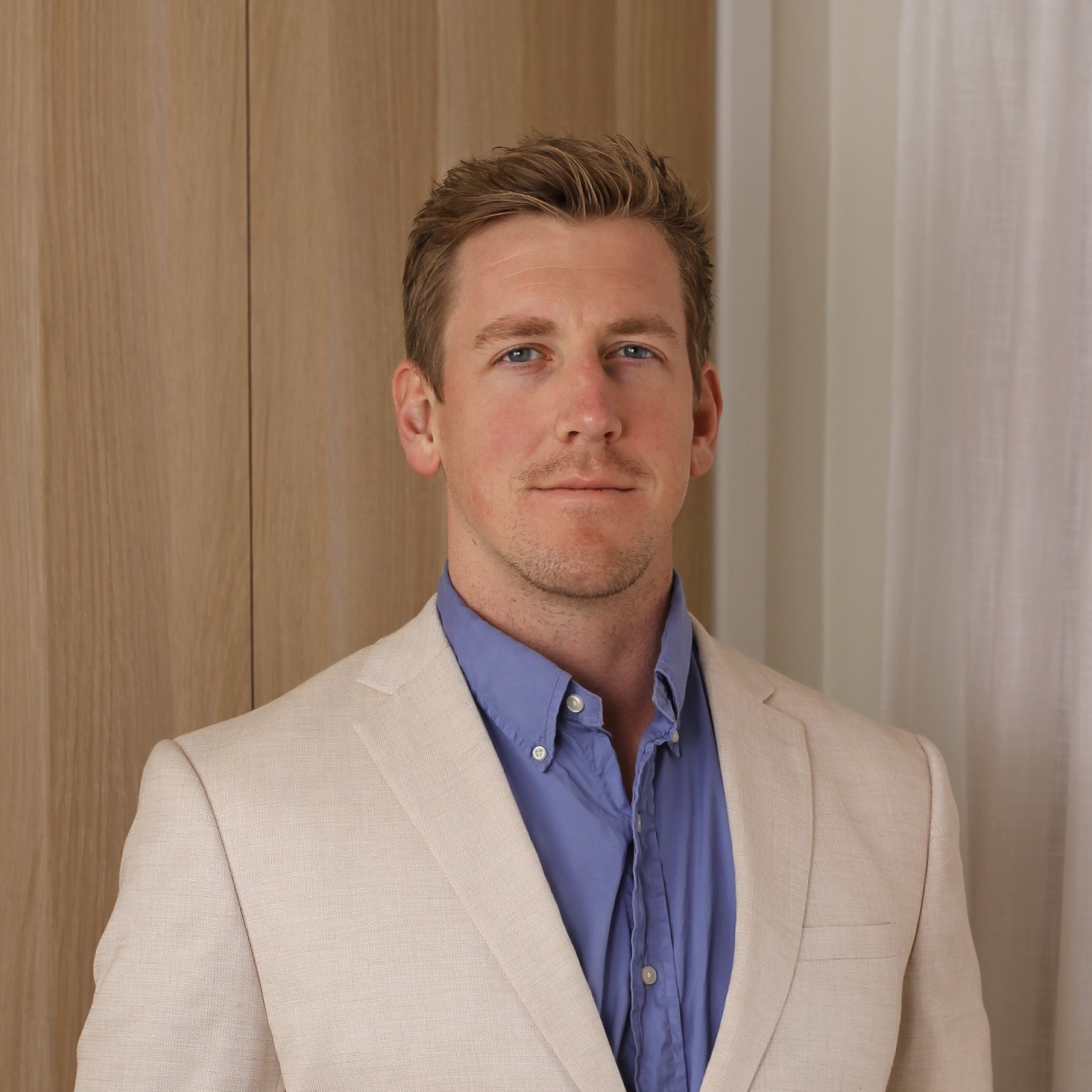 Portrait of a man with short light brown hair wearing a beige blazer and blue shirt, standing indoors against a wooden panel and white curtain background.
