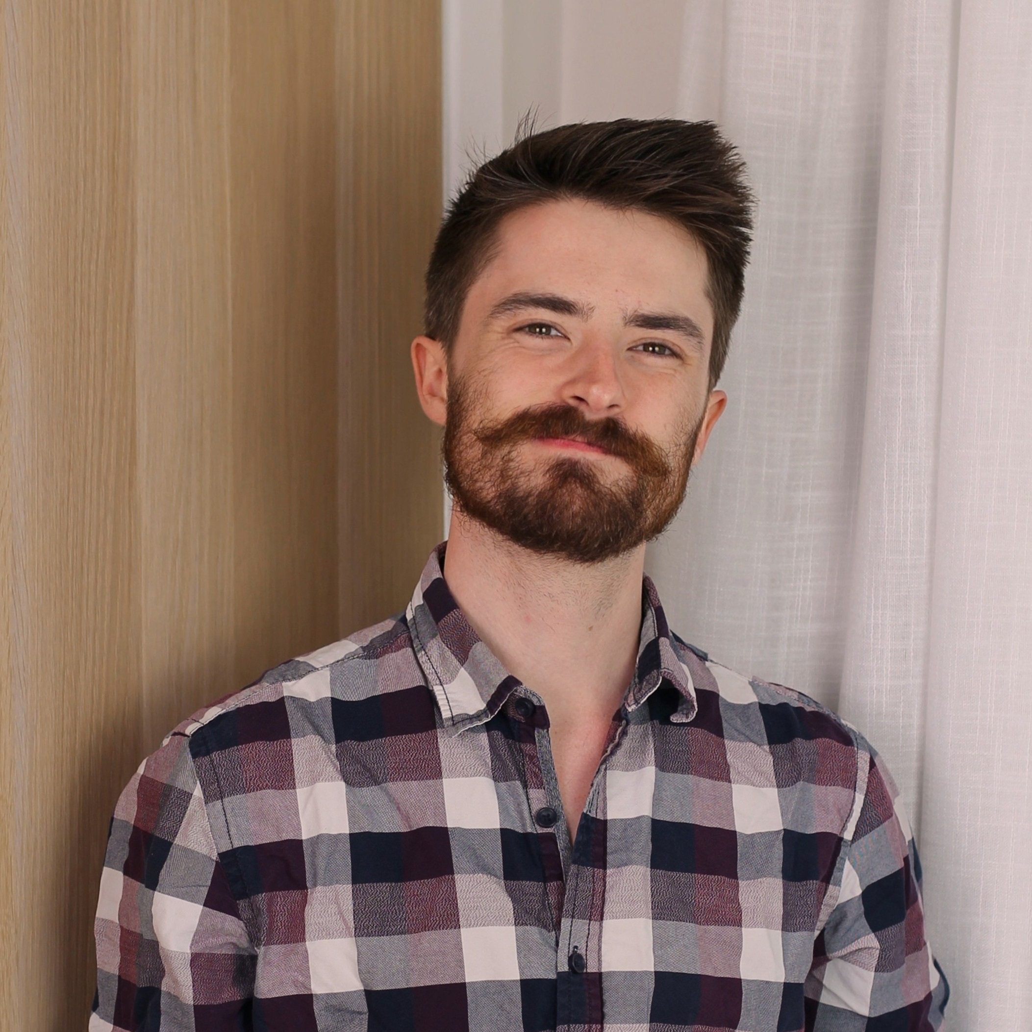 Young man with dark hair and a beard wearing a checkered shirt smiling softly against a background of wood panel and white curtain.