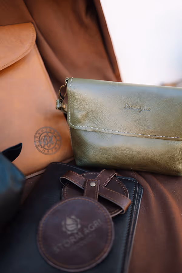 Close-up of four leather items including a green pouch with 'Blooming Nice' embossed, a brown bag with a circular logo, a black leather notebook, and a round brown leather tag labeled 'Storm Agri'.