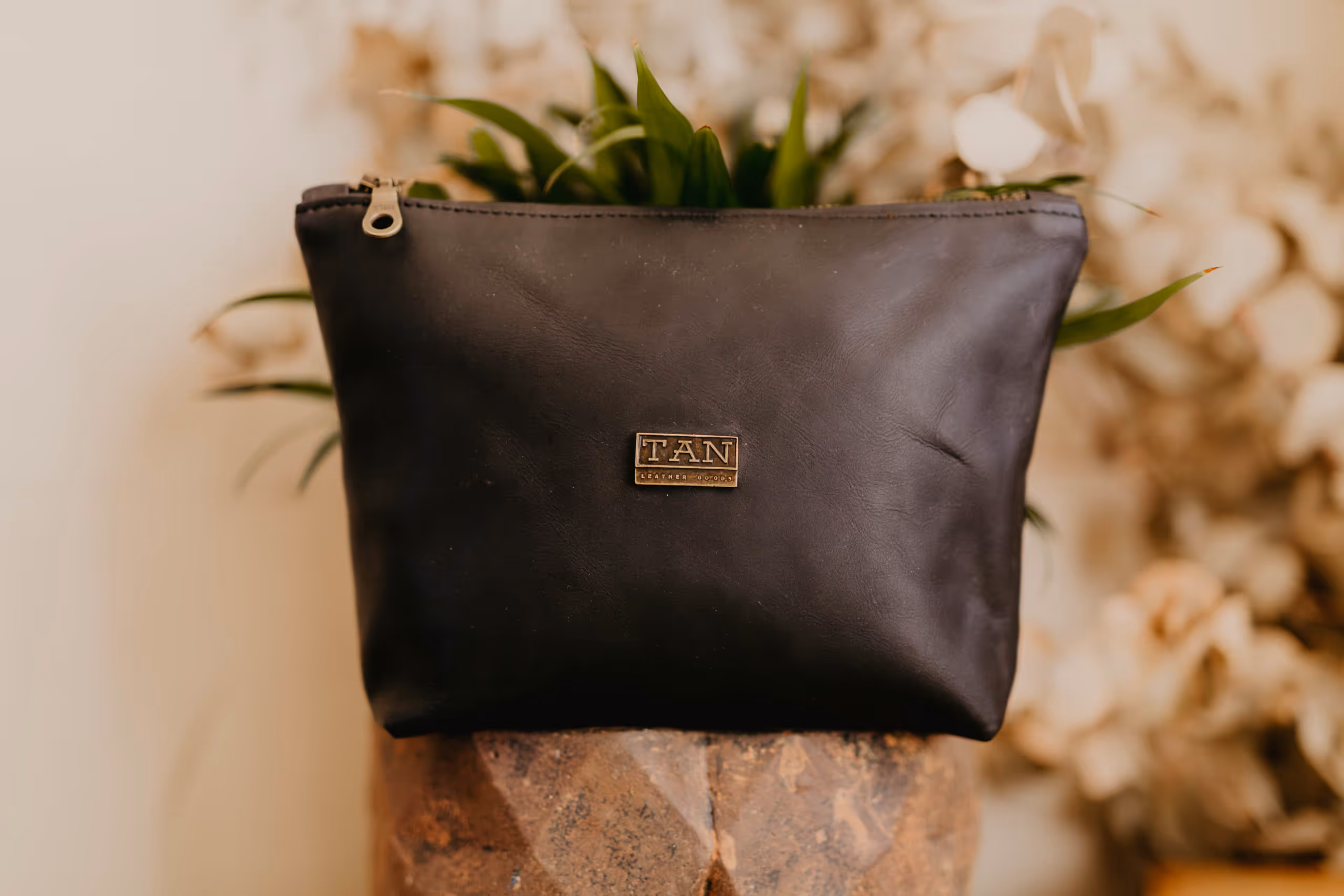 Black leather makeup bag with a metal TAN logo, positioned on a textured stone surface with green plants and blurred flowers in the background.