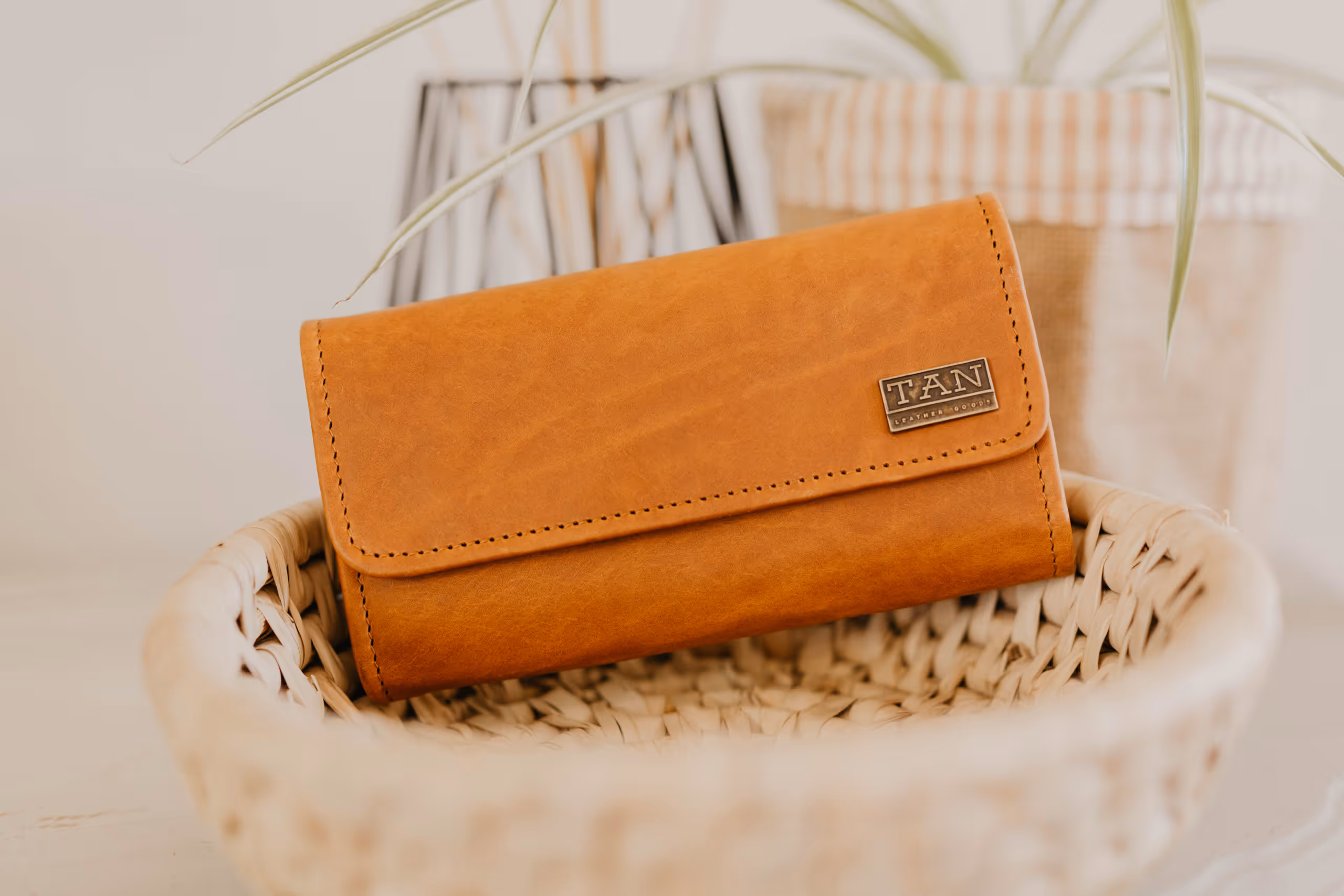 Tan leather wallet with stitched edges resting in a woven basket with a blurred plant background.