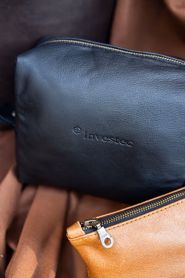 Black leather toiletry bag embossed with 'Investec' logo next to a tan leather bag with a visible zipper.