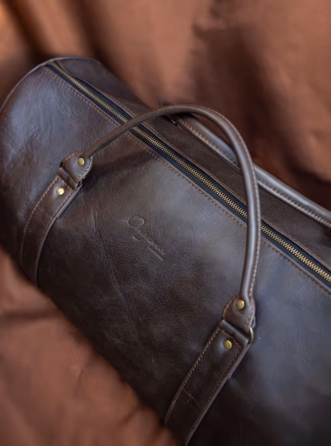 Dark brown leather duffle bag with handles and a zipper, placed on a matching brown fabric background.