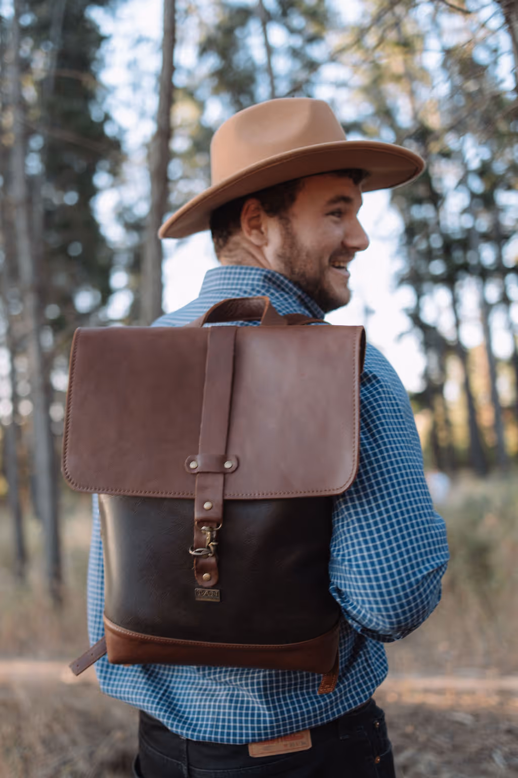 Smiling man wearing a brown hat and blue checkered shirt carrying a brown leather backpack in a forest.