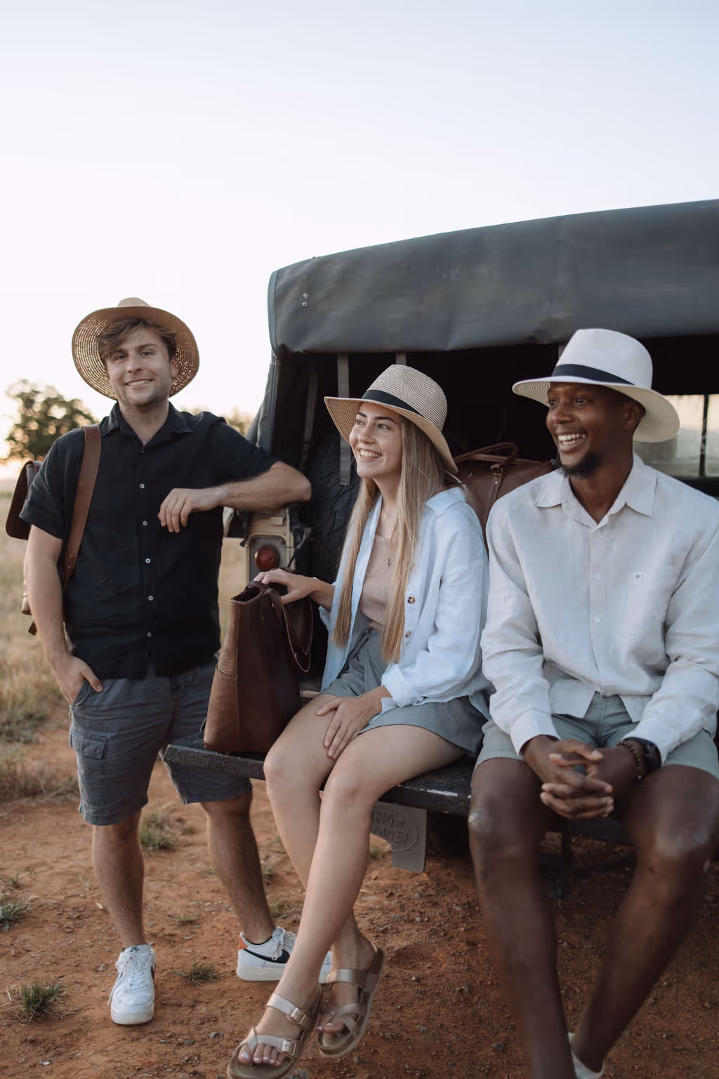 Three friends wearing hats smiling and relaxing beside an off-road vehicle in a dry outdoor setting.