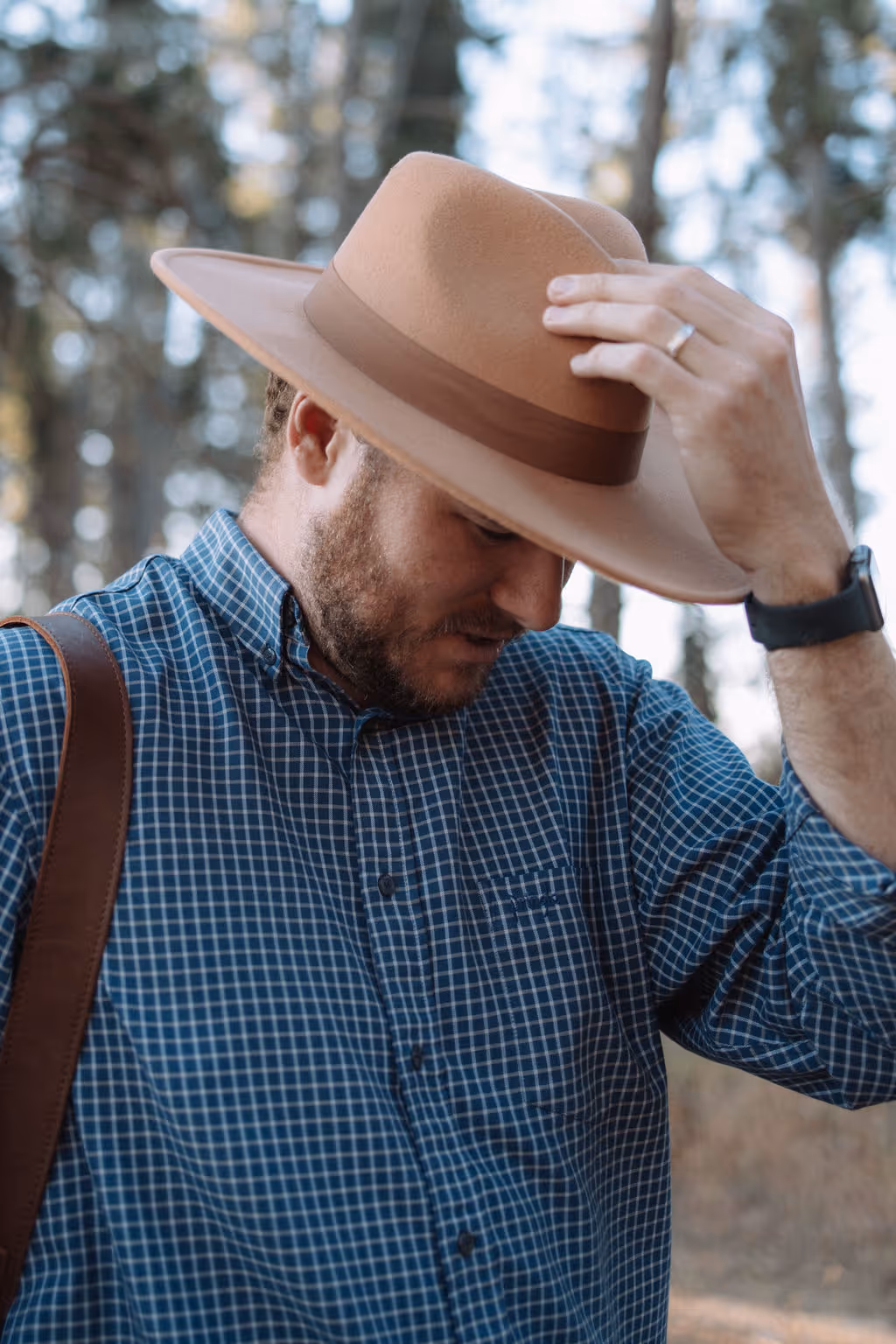 Man wearing a tan wide-brimmed hat and blue checkered shirt, adjusting his hat outdoors with blurred trees in the background.