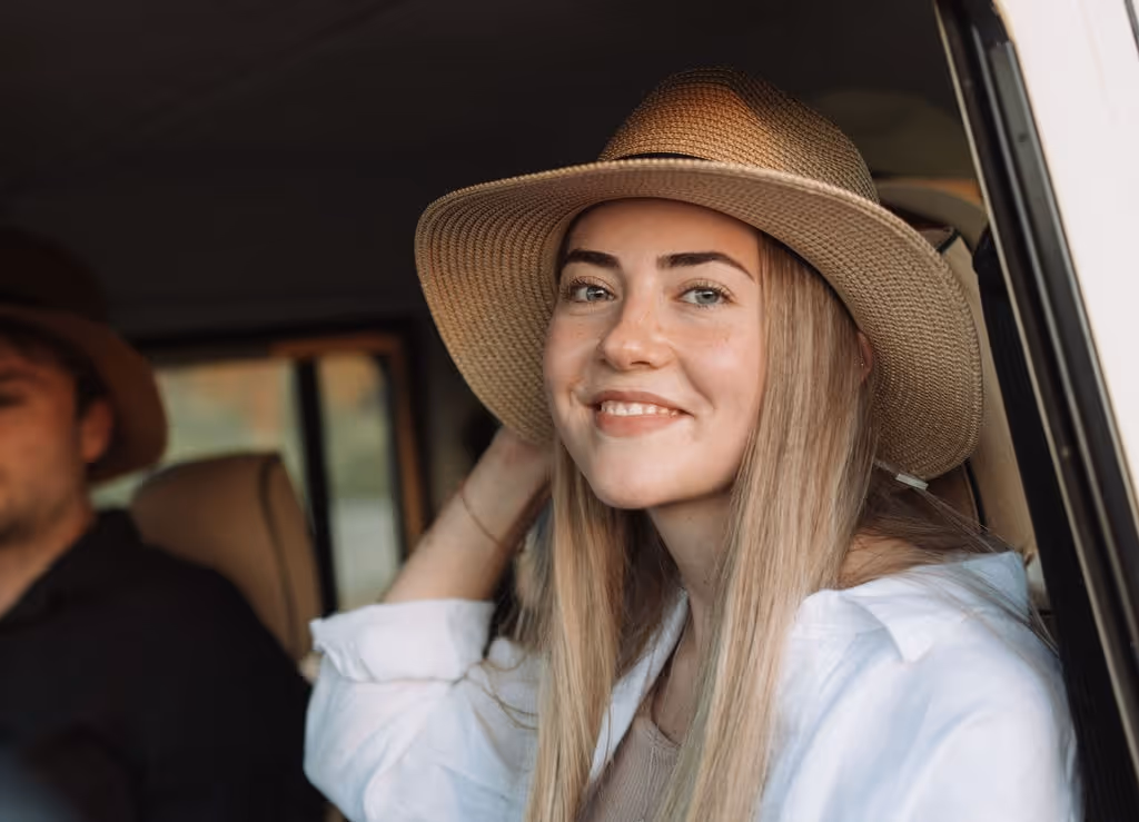 Smiling young woman with long blonde hair wearing a straw hat and white shirt sitting in a car.