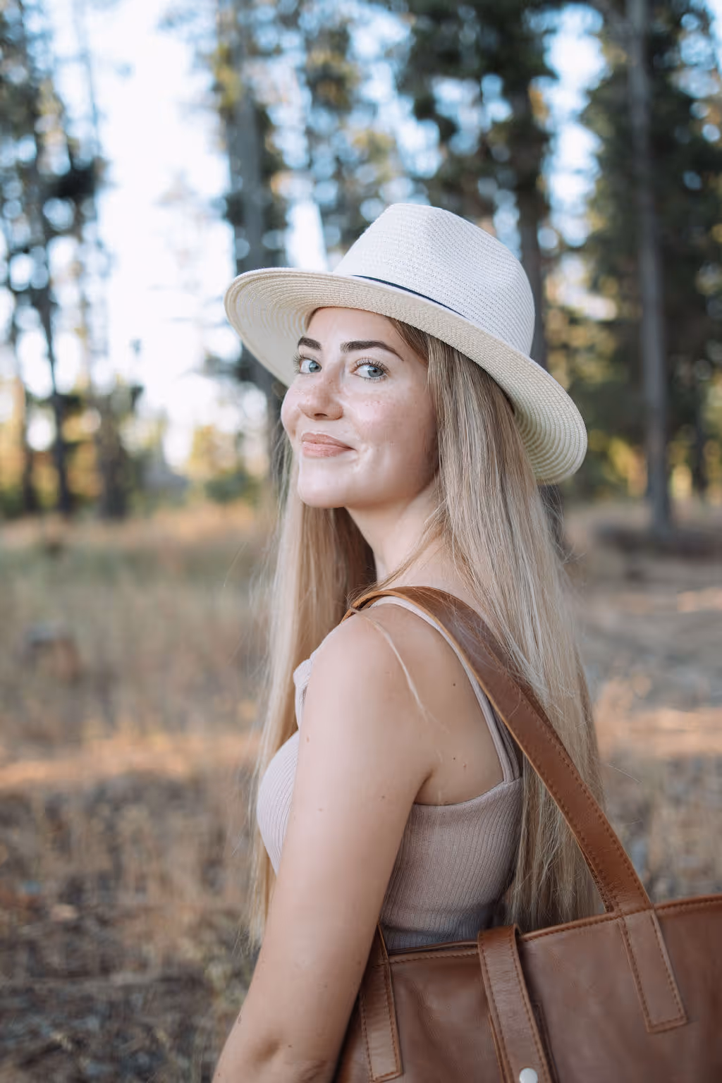 Young woman with long blonde hair wearing a white hat and carrying a brown leather bag outdoors in a wooded area.