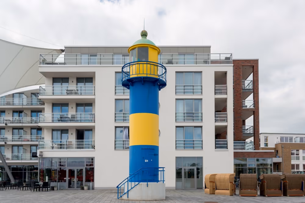 Bunter Leuchtturm mit blauen und gelben Streifen vor einem modernen weißen Gebäude mit Balkonen.