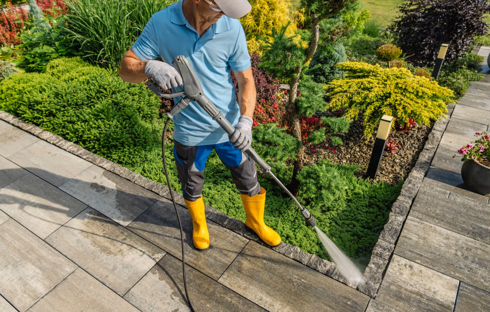 Mann in gelben Gummistiefeln und blauer Kleidung reinigt mit Hochdruckreiniger eine gepflasterte Terrasse neben einem bunten Garten.