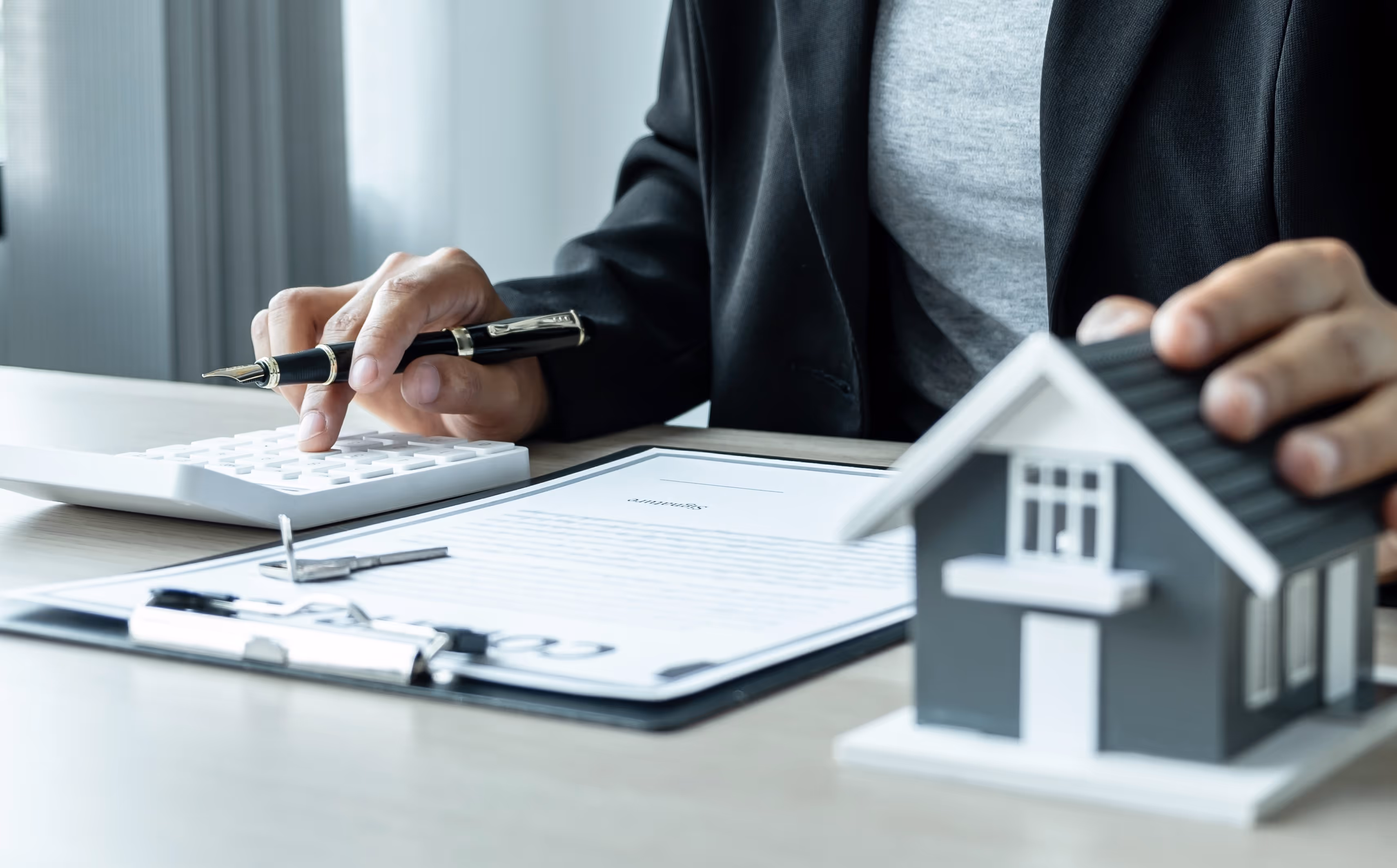 Person berechnet mit Taschenrechner und hält einen Stift, während er ein Hausmodell berührt und einen Vertragsdokument auf dem Tisch hat.