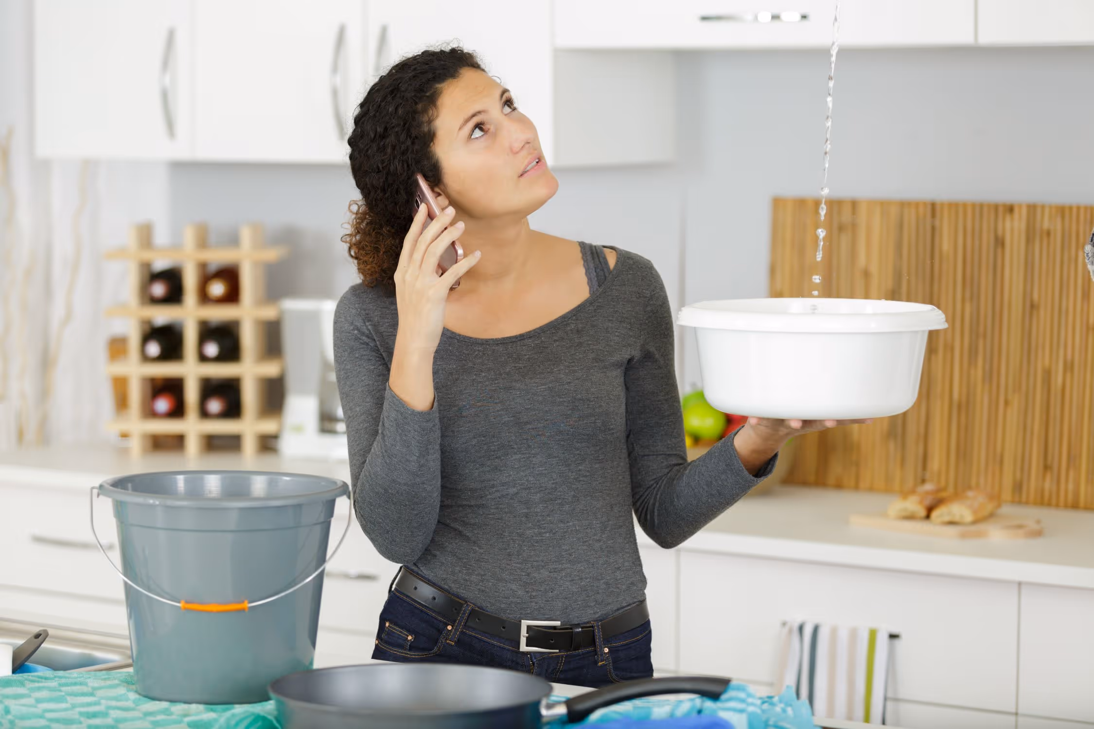 Frau in der Küche hält einen Eimer unter einer undichten Decke, während sie telefoniert.