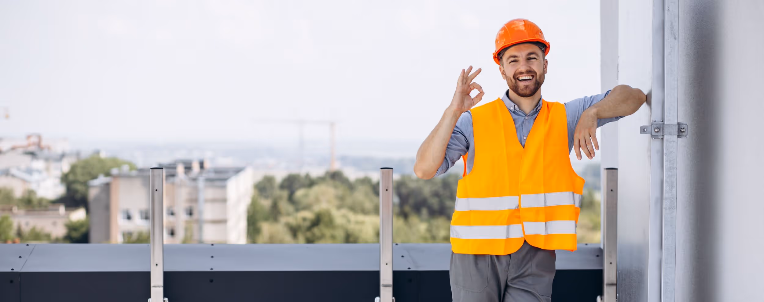 Lächelnder Bauarbeiter mit oranger Sicherheitsweste und Helm zeigt das Okay-Zeichen auf einer Baustelle.