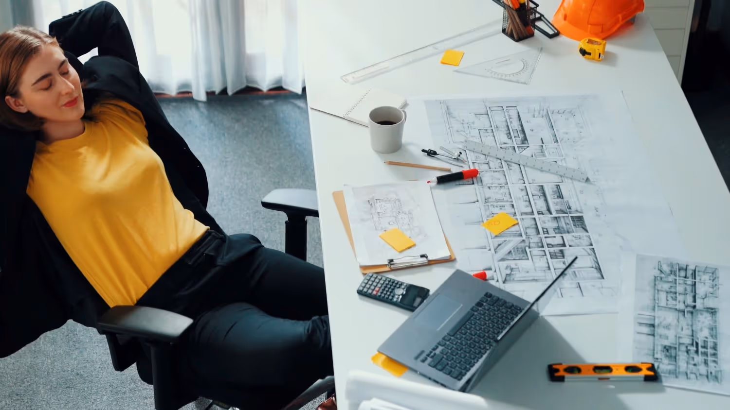 Frau entspannt sich im Bürostuhl neben einem Schreibtisch mit Computer, Bauplänen, Taschenrechner und Helm.