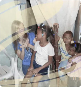 A woman with blonde hair sitting and talking with three children, one girl sits on her lap and two children sit nearby on chairs.