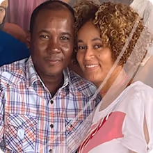 Smiling couple posing closely together, with the man wearing a plaid shirt and the woman in a white top with curly hair.