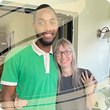 A man in a green polo shirt, Hendrix, with white trim smiling with his arm around a woman wearing glasses and a black shirt inside a room.