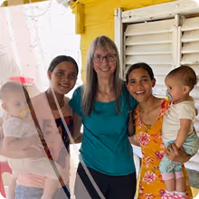 4 women smiling, with two holding  babies, Lilly bear and Lilly Anna, on each side, standing in front of a yellow wall and white window shutters.