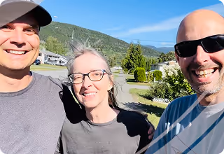 Three smiling adults, Ryan, Vivian and Shane, outdoors on a sunny day with hills and houses in the background. 