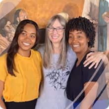 Three smiling women standing close together with their arms around each other, with a mural painting in the background.