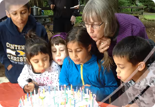 A group of children and an older woman gathered around a birthday cake with lit candles, preparing to blow them out.