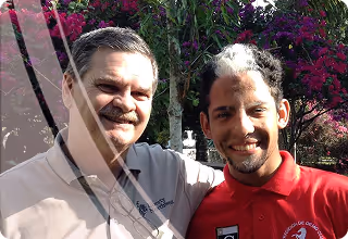 Two men smiling outdoors with trees and pink flowers in the background.
