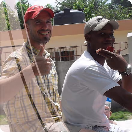 Two men sitting on a motorbike, one smiling and giving a thumbs up, the other making a hand gesture while wearing caps.