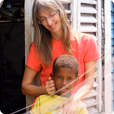 Smiling woman with glasses in a red shirt embraces boy in a yellow basketball shirt in front of a shuttered window.