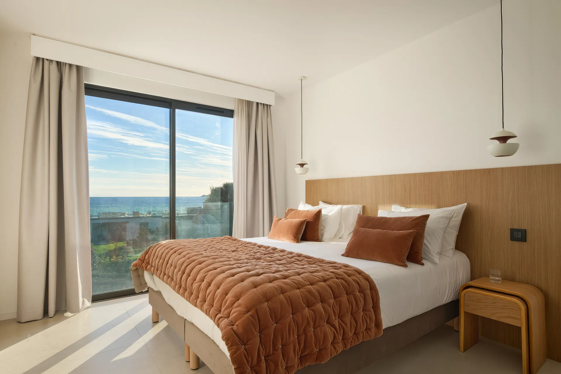 Chambre 8  moderne avec grand lit double, couverture et coussins couleur terre cuite, vue sur la mer par une baie vitrée.