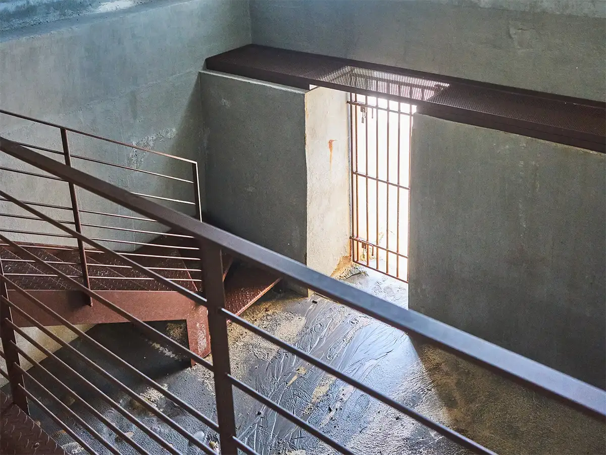 Escalier métallique avec rambarde et porte en fer forgé laissant passer la lumière dans une pièce en béton, passage du monte cristo.  