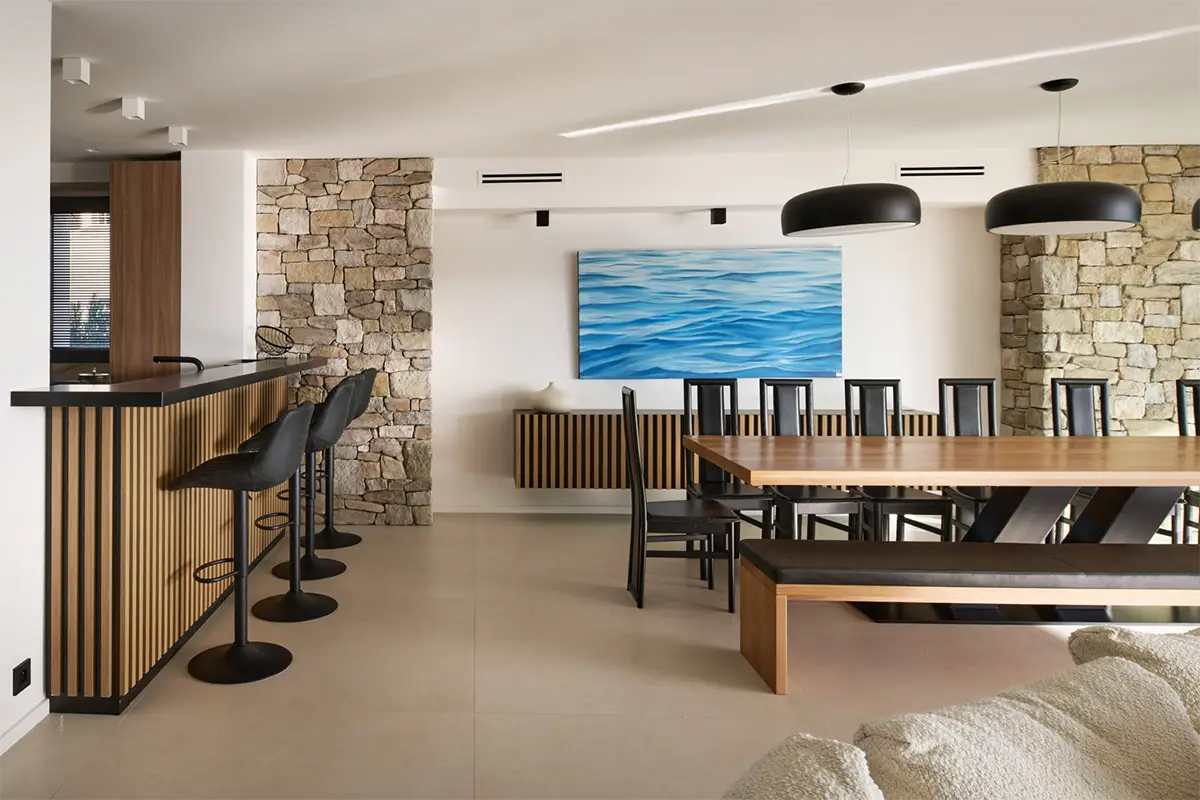 Salle à manger la villa l'île verte avec table en bois, banc, chaises noires, bar avec tabourets et mur en pierre décoré. La Ciotat