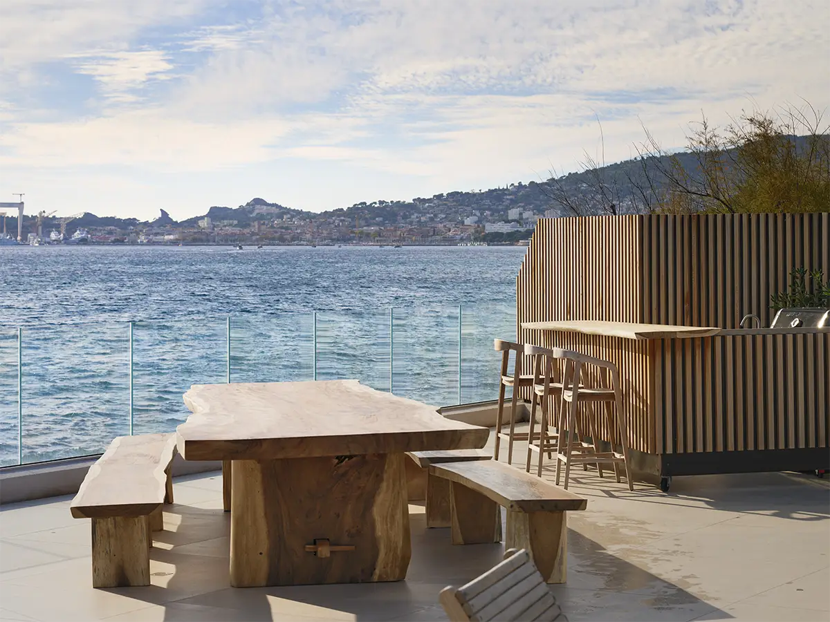 bar lounge du pavillon de la villa de luxe L’Île Verte avec coin repas extérieur, table et bancs en bois face à une balustrade en verre offrant une vue panoramique sur la mer et les collines méditerranéennes à La Ciotat