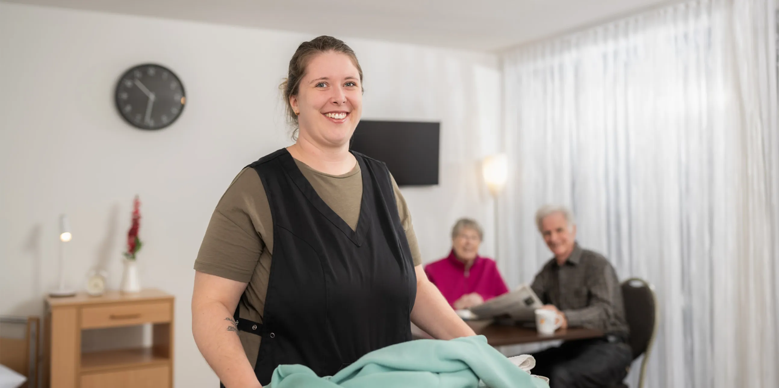 Ein älterer Mann mit Brille sitzt, während eine jüngere Frau hinter ihm steht und beide lächelnd in die Ferne blicken.