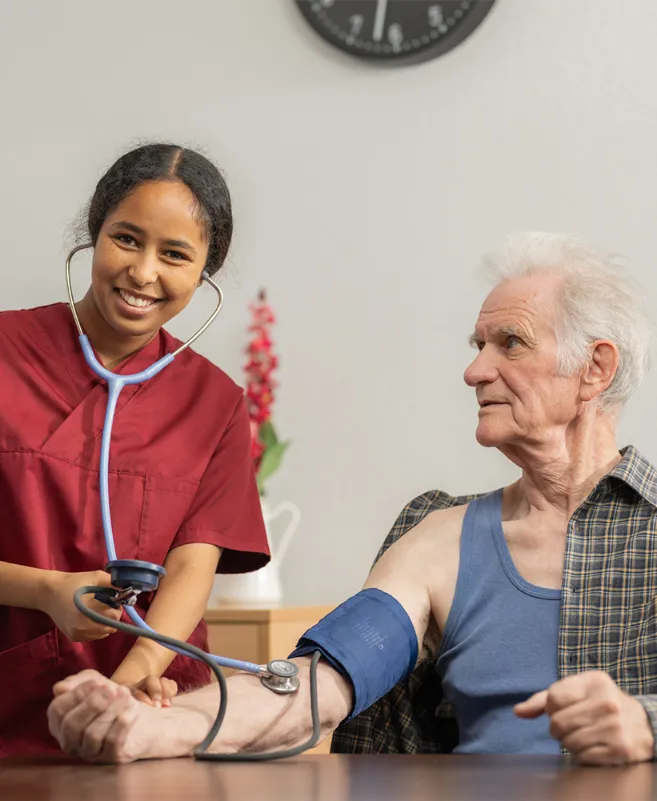 Eine Lernende lächelt in die Kamera während sie bei einem älteren Mann den Blutdruck misst.