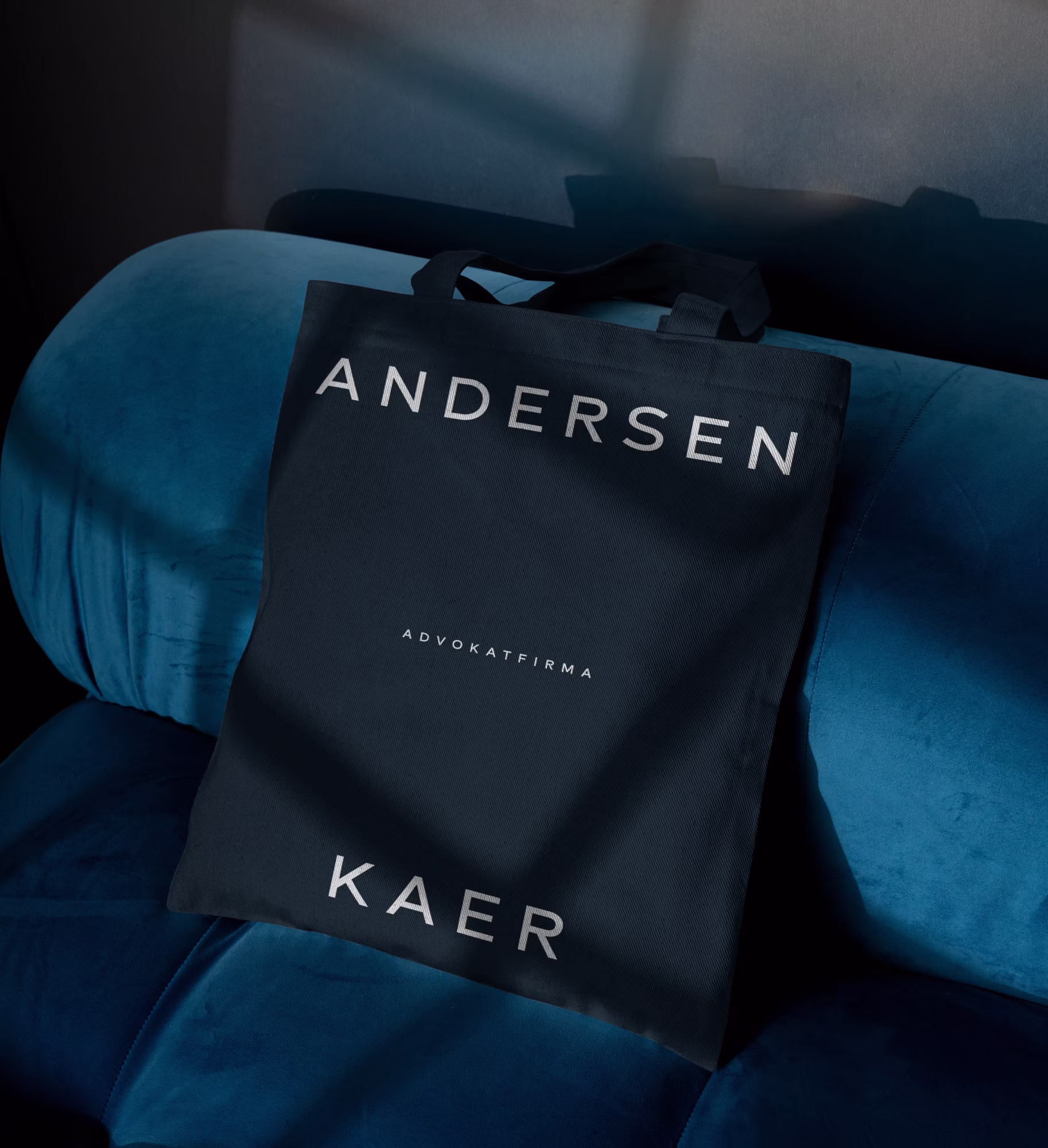 Sort mulepose med hvid tekst "ANDERSEN KAER ADVOKATFIRMA" placeret på blå velourstol.