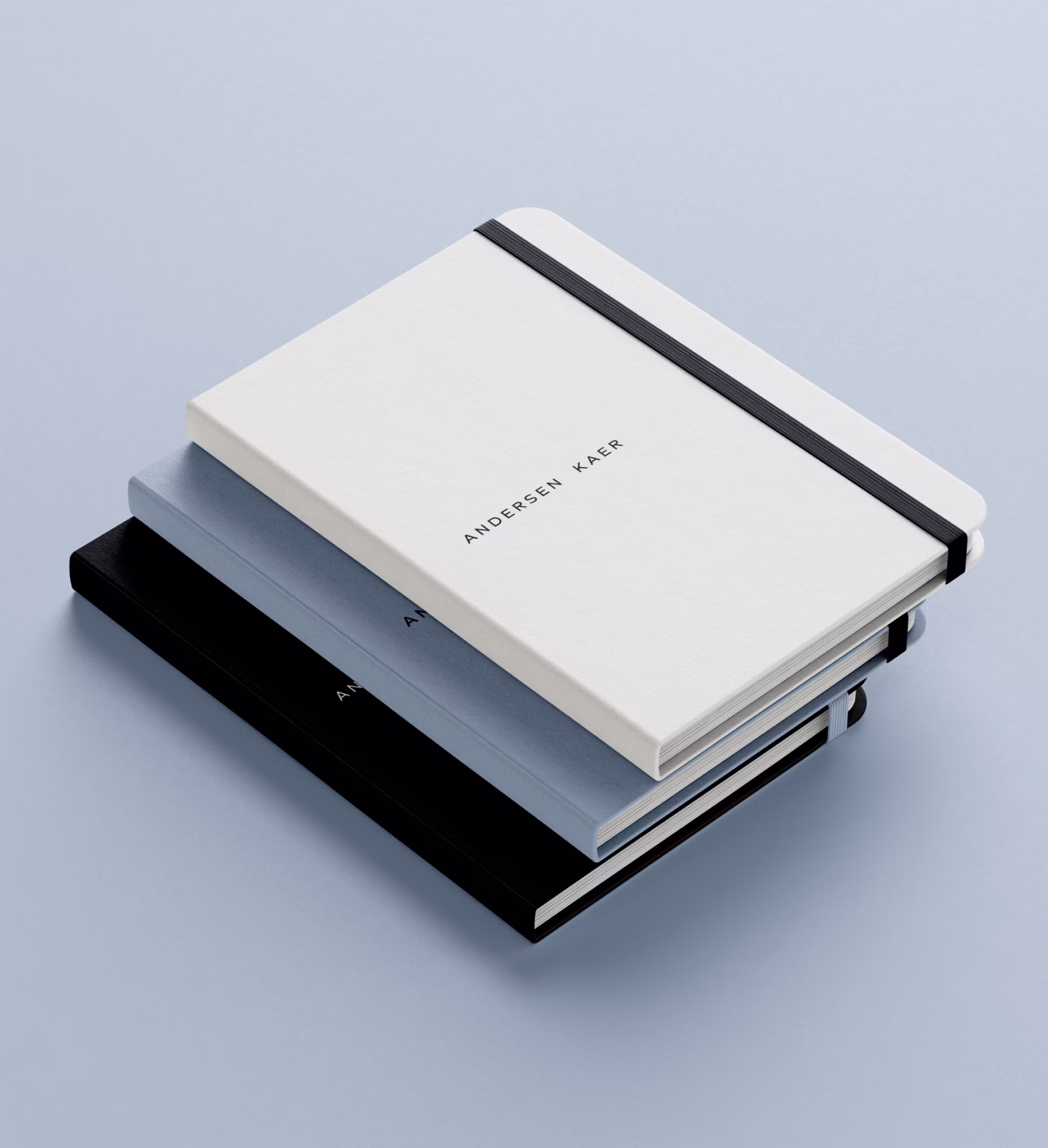 Stak af tre notesbøger i hvid, lyseblå og sort med elastikbånd på lyseblå overflade.