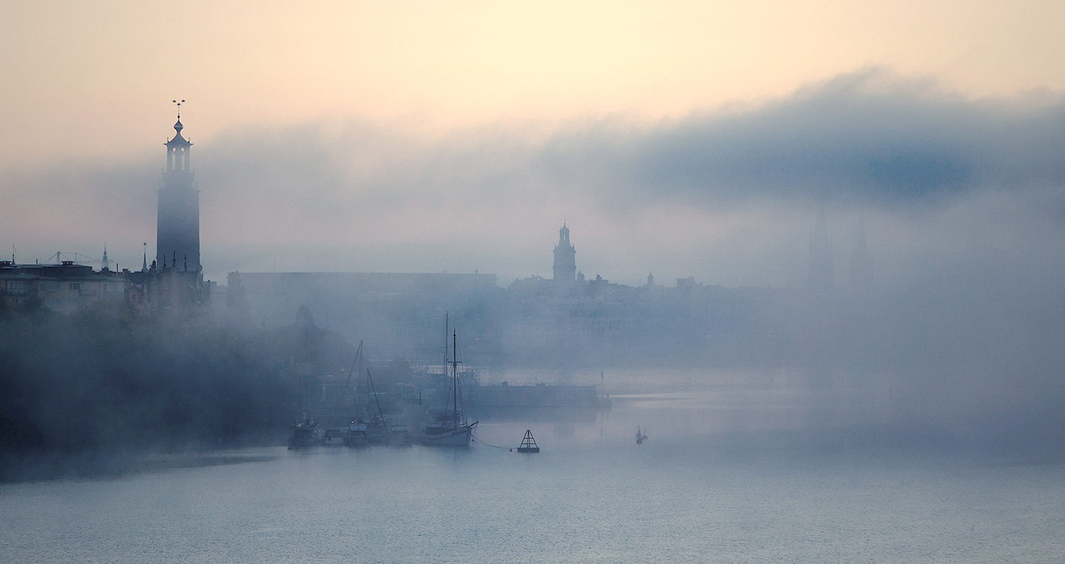 Foggy waterfront scene with silhouetted historic towers and boats partially obscured by mist at dawn.