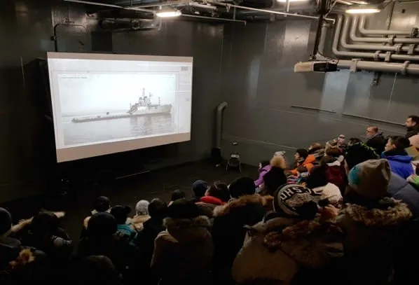 Audience wearing winter clothing watches a black and white naval ship video projected in a dark room with exposed pipes.
