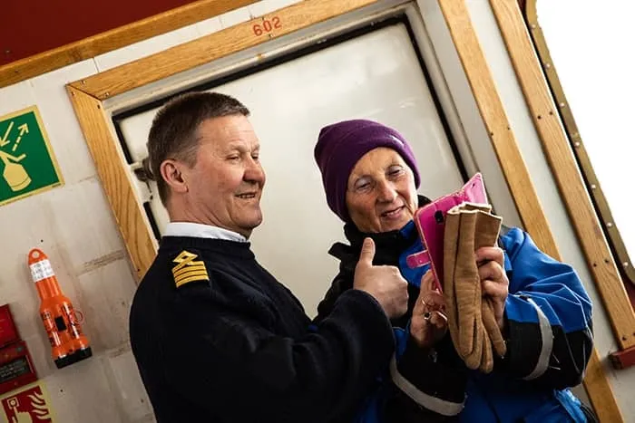 A man in a navy uniform smiles and gives a thumbs-up while standing next to a woman in a purple beanie and blue jacket holding brown gloves and a pink smartphone cover.
