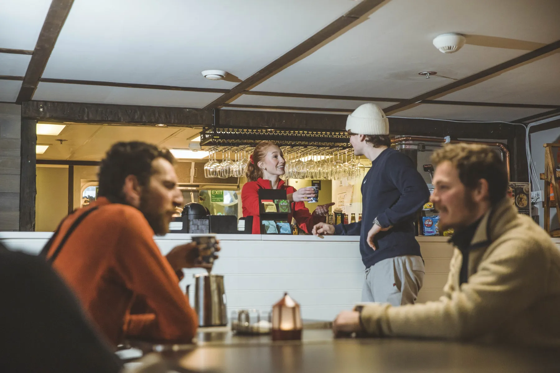 A woman in a red jacket serves a drink to a man in a white beanie across a counter while two men sit and drink in the foreground.