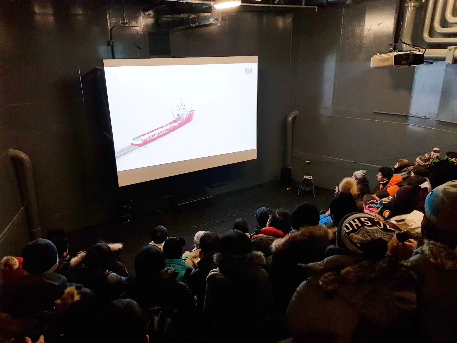 A group of people wearing winter clothing watching a movie of a red ship on a large screen in a dark room.