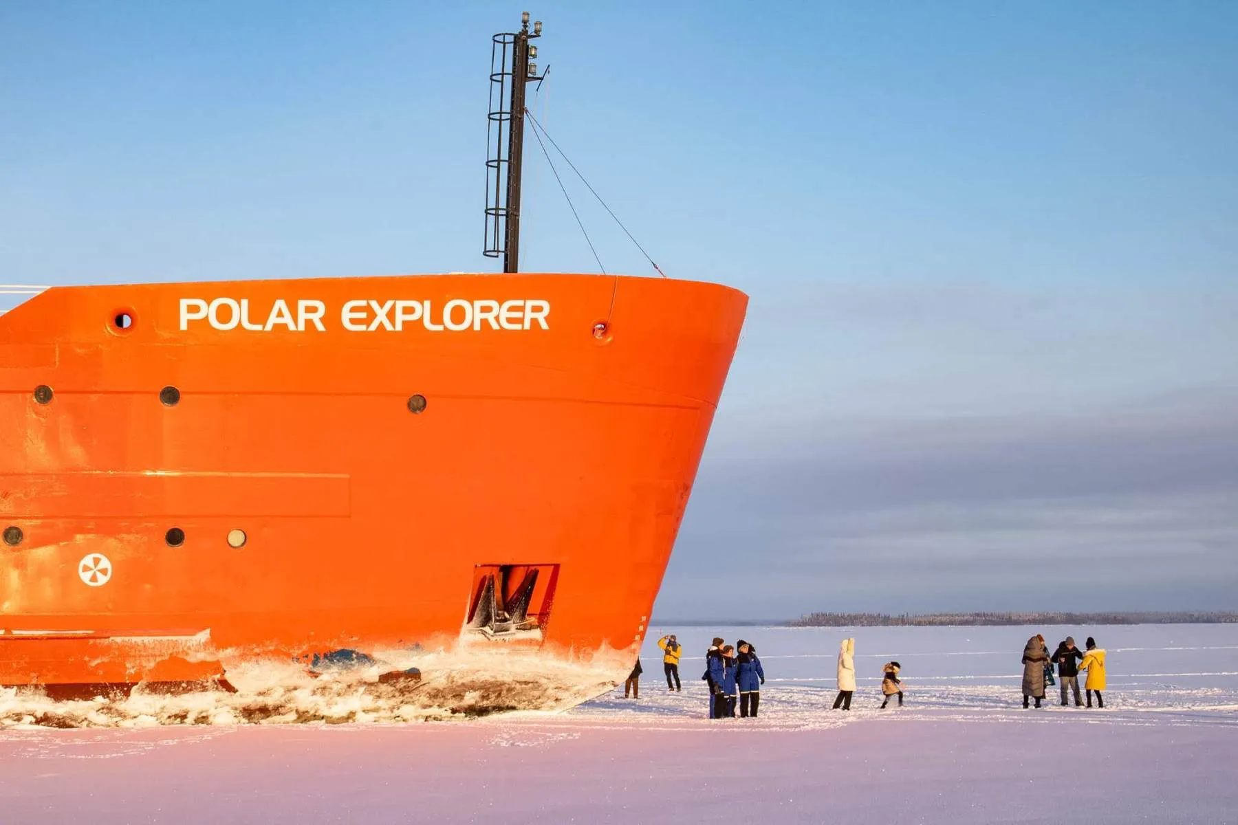 The bright orange bow of the Polar Explorer ship navigates through icy waters near a group of people standing on the snow-covered ice under a clear sky.