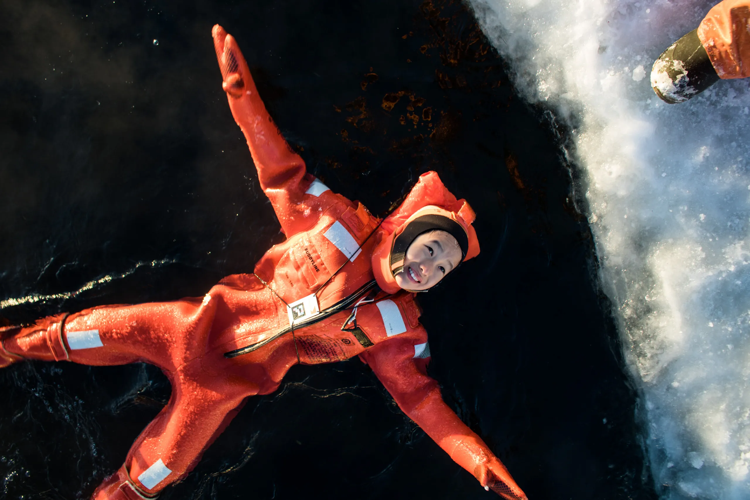 Person wearing an orange cold water survival suit floating in icy water near a chunk of ice.
