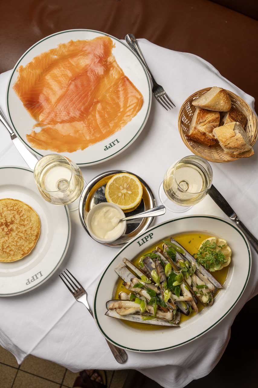 Table with plates Pillivuyt of smoked salmon, razor clams in sauce with herbs, a blini, bread basket, lemon wedge, and two glasses of white wine at iconic Brasserie Lipp.