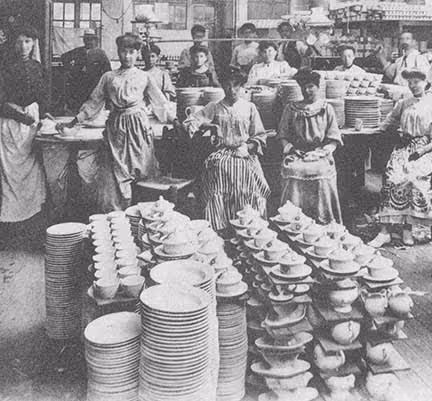 Photo historique en noir et blanc de la manufacture Pillivuyt montrant plusieurs femmes en robes longues dans une usine préparant et empilant de la vaisselle en porcelaine.
