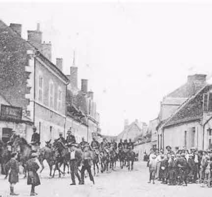 Photo de la ville de Mehun-sur-Yèvre en noir et blanc. Une troupe de cavaliers en uniforme passant dans une rue bordée de maisons, observée par un groupe de civils, hommes, femmes et enfants.
