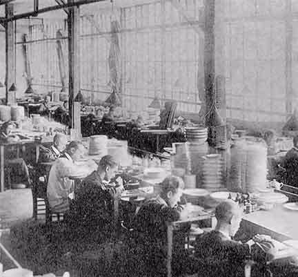 Photo historique Pillivuyt. Des ouvriers Pillivuyt assis à une longue table dans un atelier, assemblant ou inspectant des objets, avec des piles de produits autour d'eux.