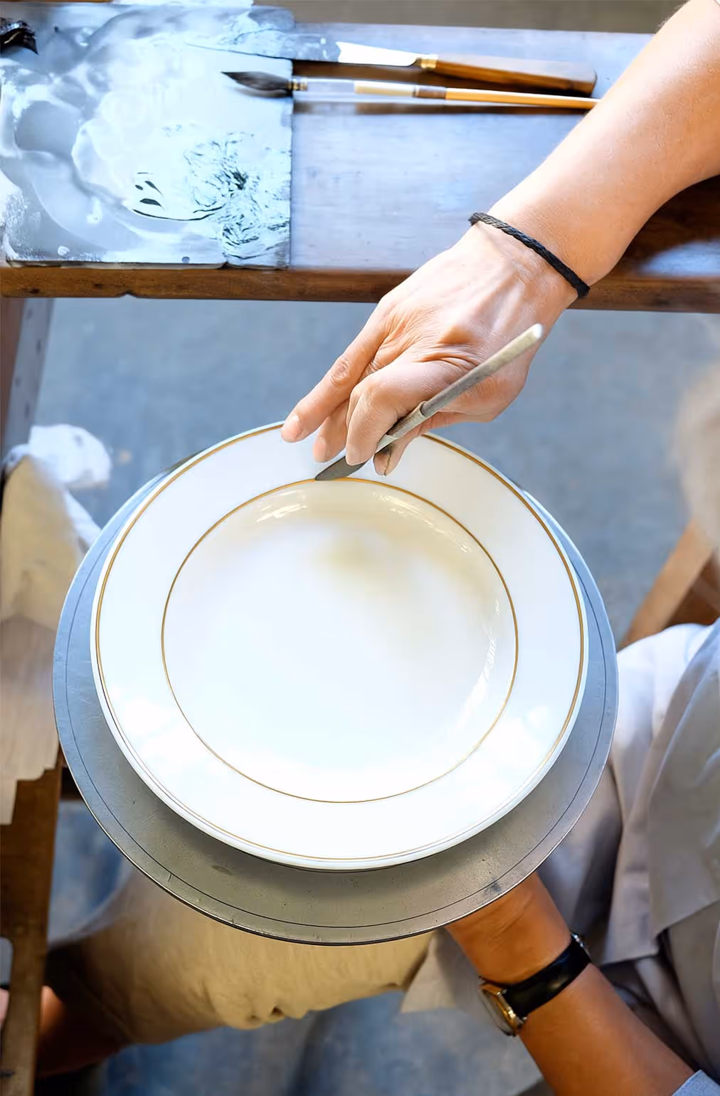 Pillivuyt artisan holding a white ceramic plate with gold rim on a pottery wheel, sculpting its edge with a shaping tool.